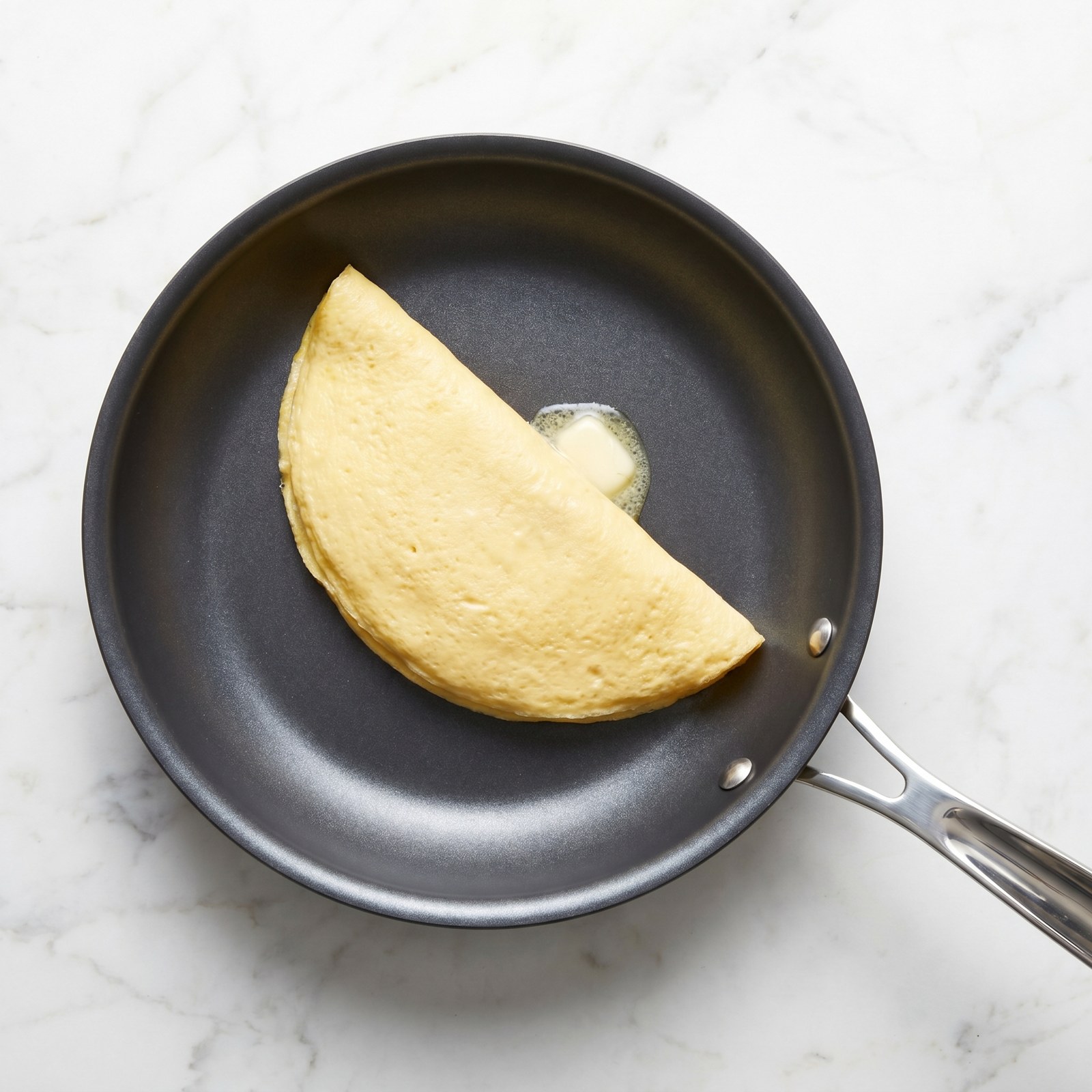 8-inch nonstick omelette pan with a folded golden omelette and melting butter, overhead on marble
