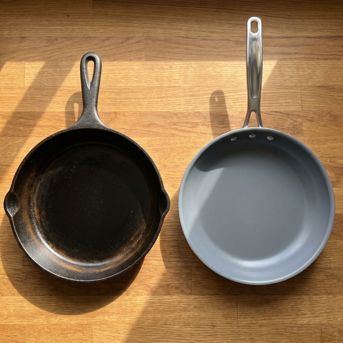 Dark seasoned cast iron skillet beside a grey nonstick frying pan on a wooden counter in natural light