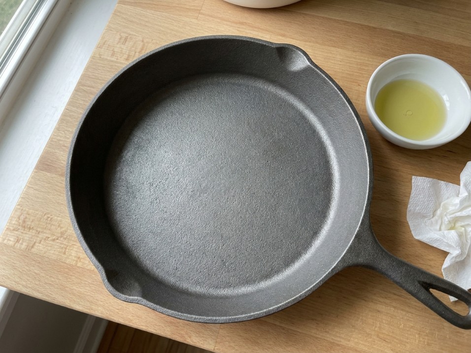New cast iron skillet next to a small dish of oil and a folded paper towel, overhead shot on a wood surface