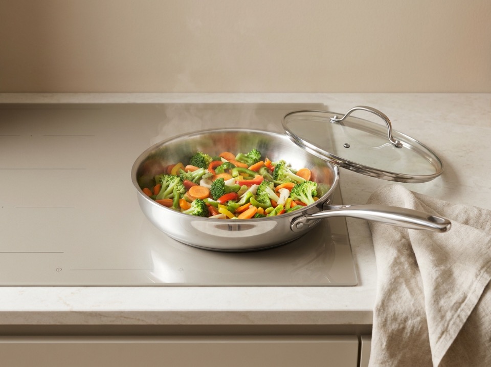 A stainless steel frying pan on a stovetop with vegetables steaming inside and a glass lid resting beside it