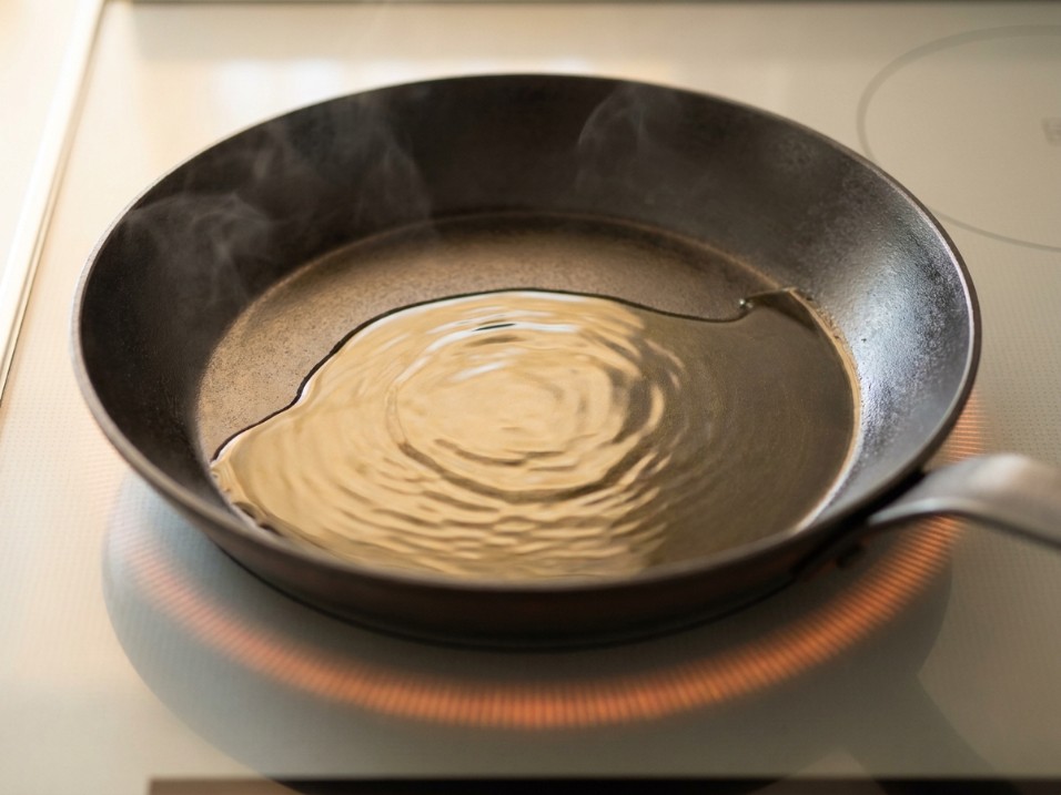 Carbon steel pan on an electric glass-top stove with oil visibly rippling, steam rising from the surface